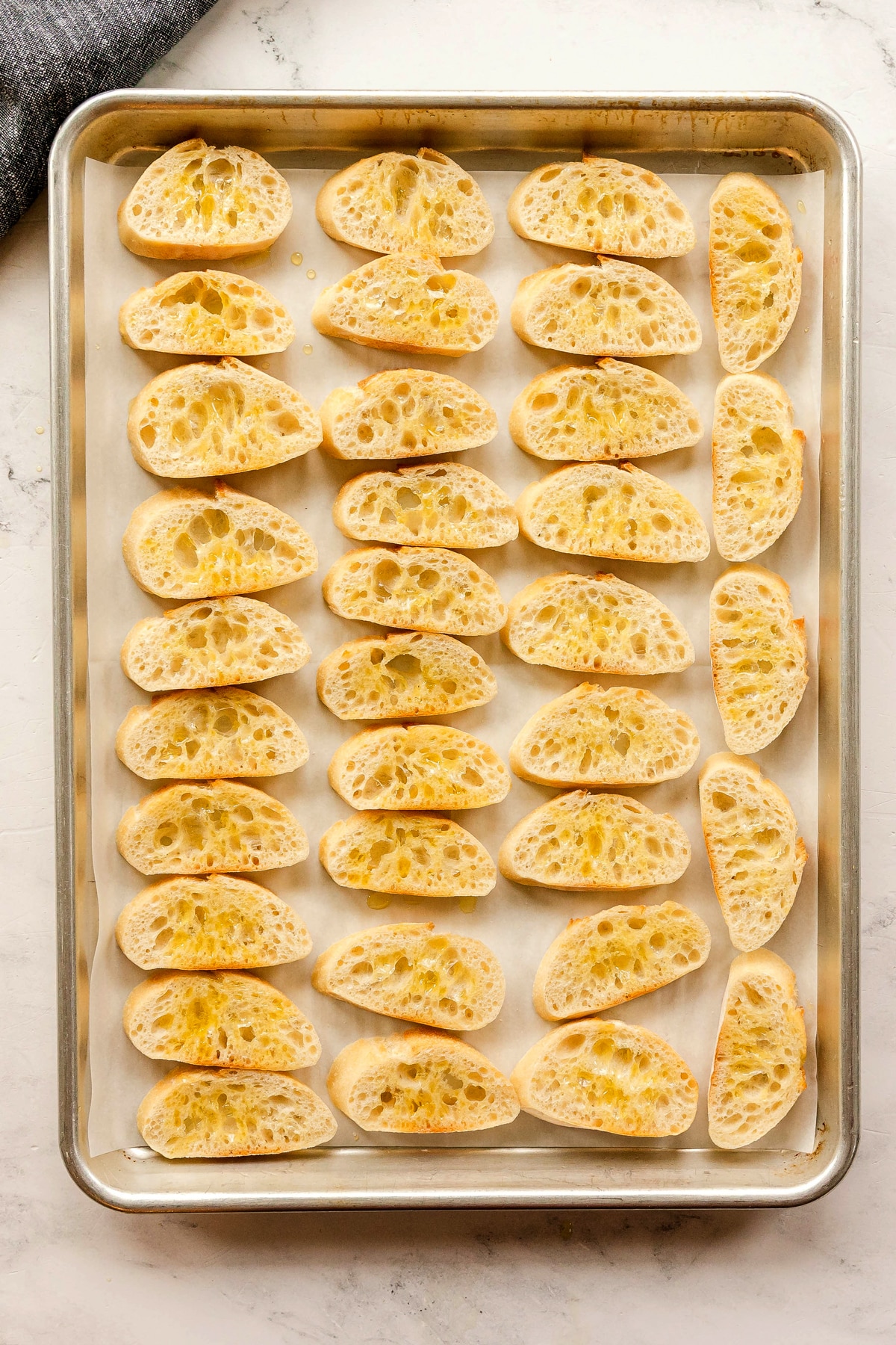 A baking sheet lined with parchment paper holds 28 slices of baguette arranged in rows, each slice drizzled lightly with olive oil.