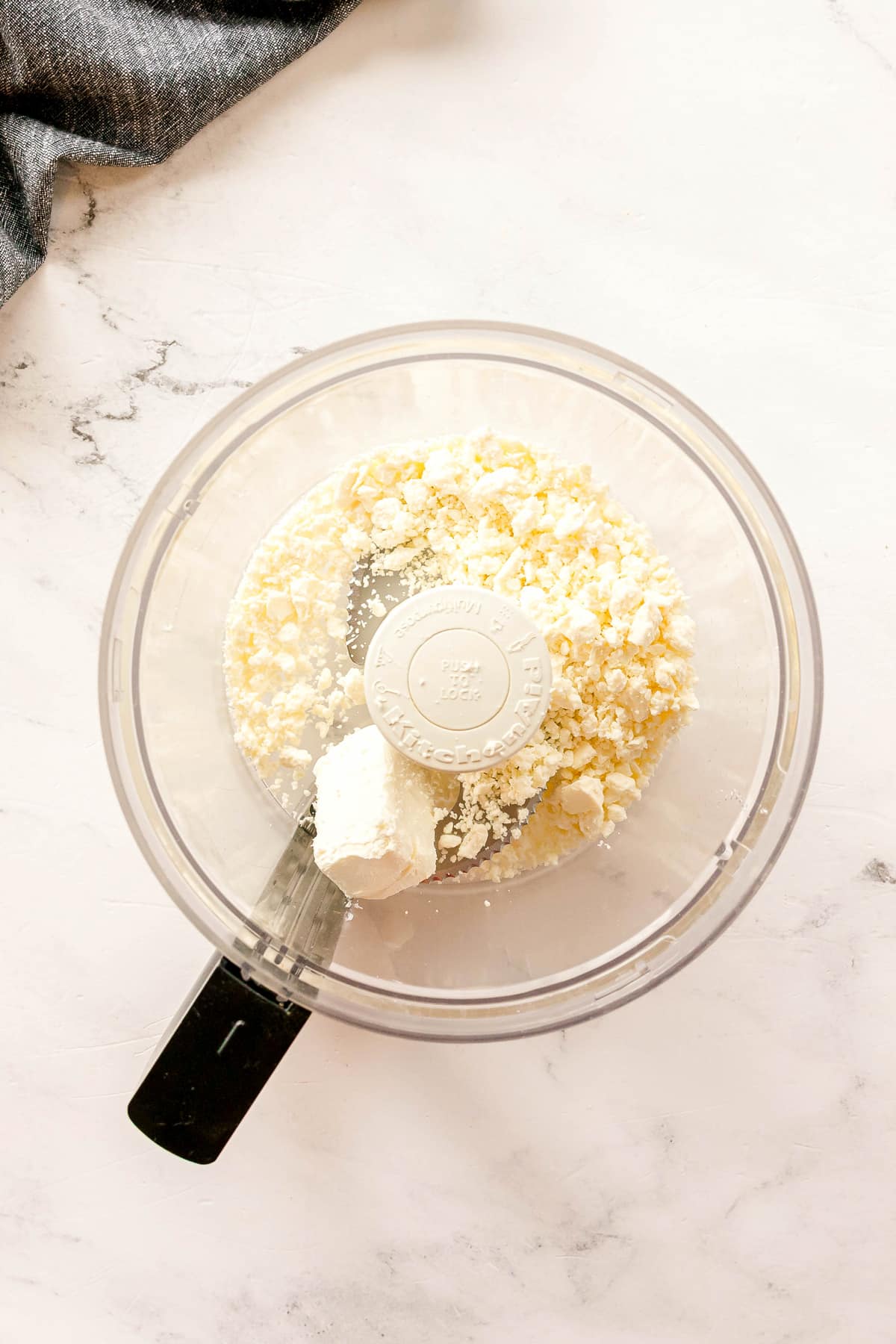 A food processor bowl containing feta and cream cheese on a white countertop, viewed from above for Whipped Feta Tomato Crostini recipe.
