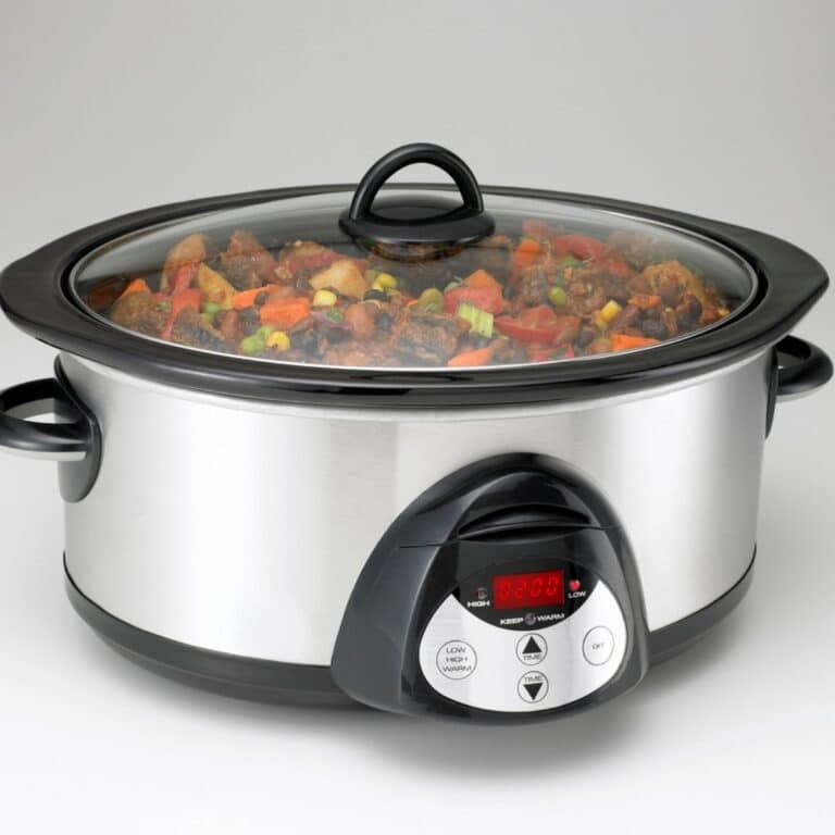 A stainless steel slow cooker filled with stew, featuring vegetables and meat, sits on a gray surface. The digital display shows a cooking time and control buttons are visible.