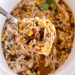 A spoonful of Slow Cooker Southwestern Chicken—featuring shredded chicken, corn, black beans, and tomatoes in a savory broth—is held above a bowl containing the same hearty mixture.