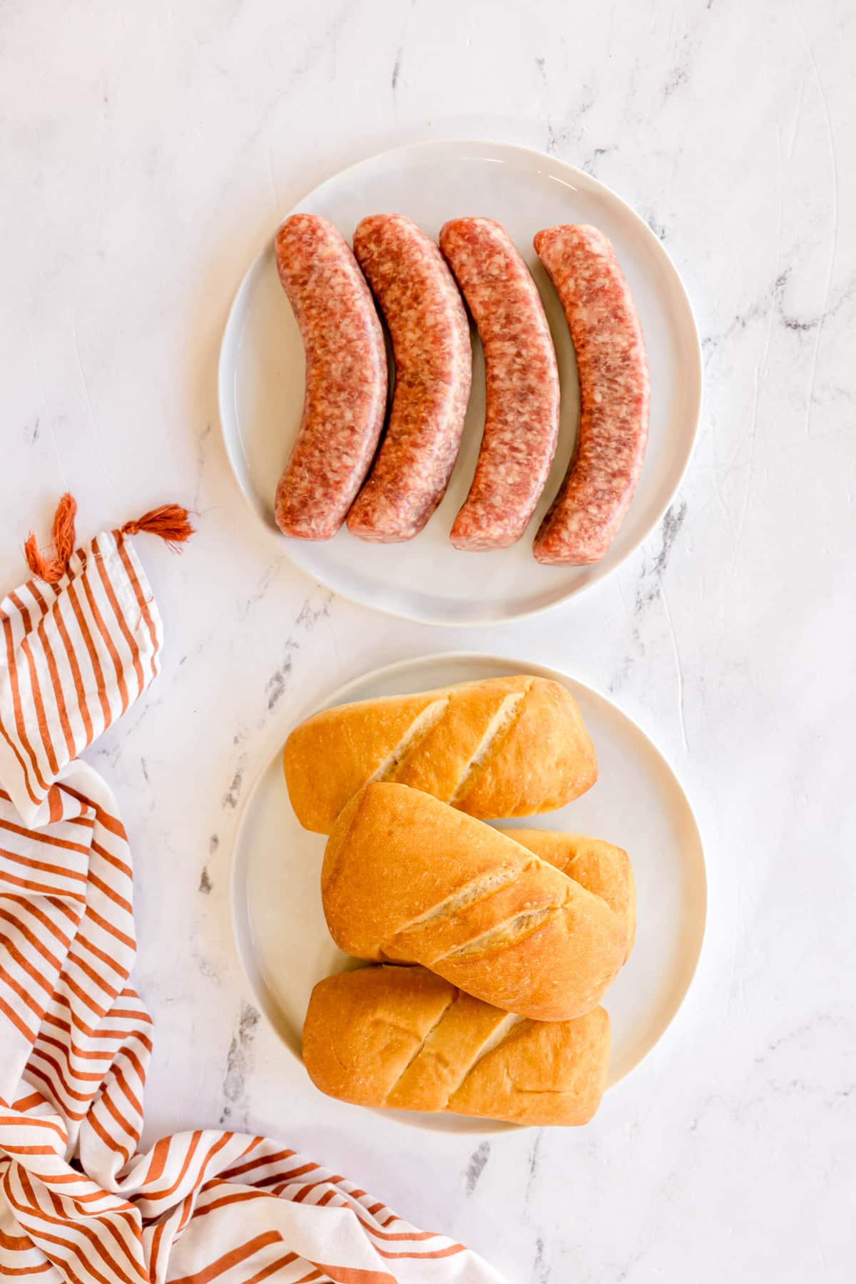 Ingredients Air Fryer Brats include uncooked bratwursts, brat or sausage buns on white plate and striped cloth to the left on a white marble surface.