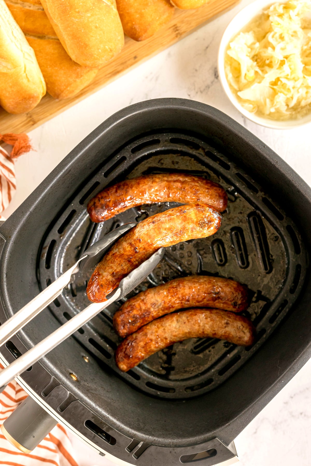 A tong holds one of Air Fryer Brats. Sides of bread rolls and sauerkraut are nearby on a white surface.