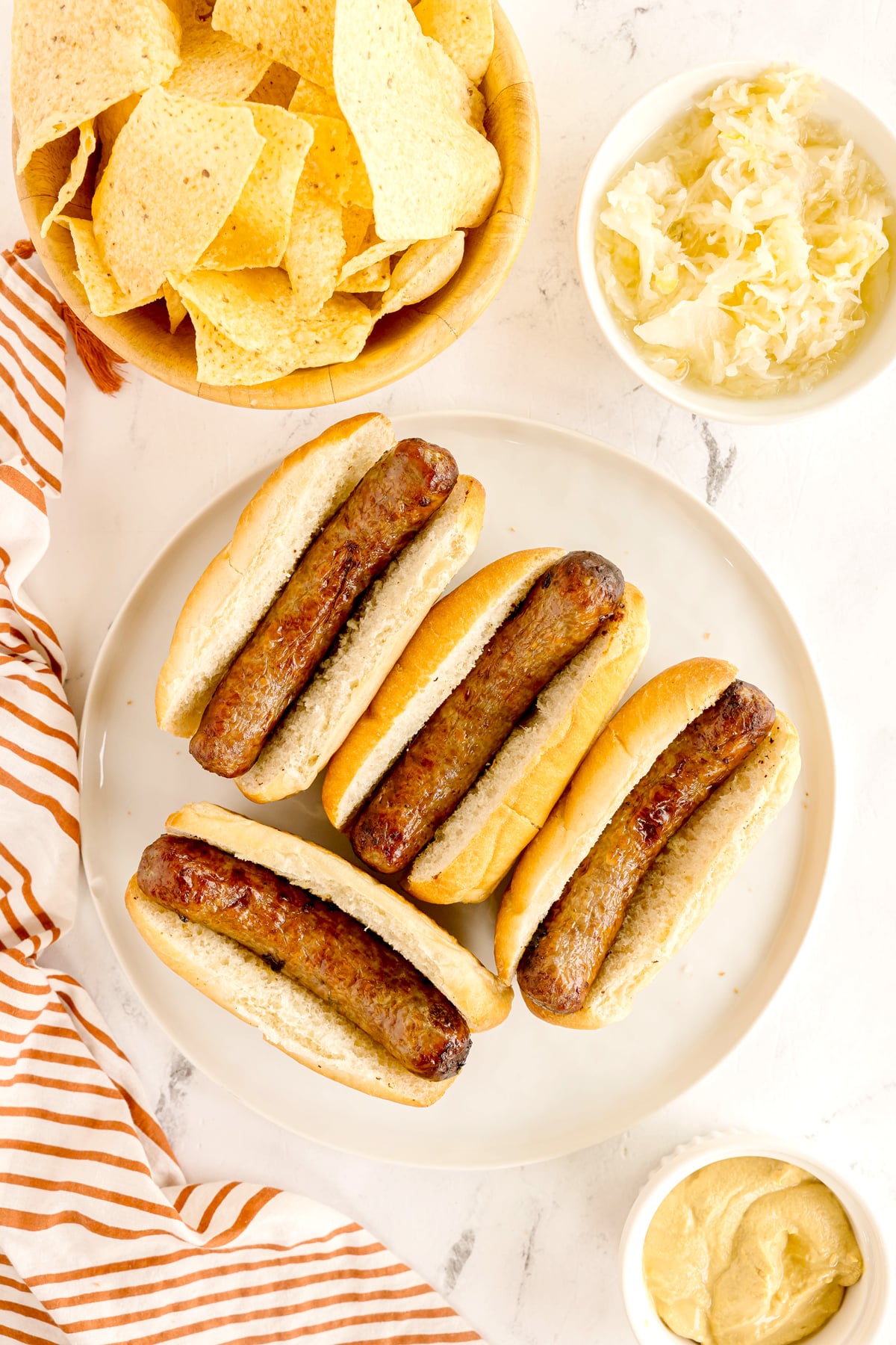 A plate with four Air Fryer Brats with buns, surrounded by bowls of tortilla chips, sauerkraut, and mustard on a white surface.