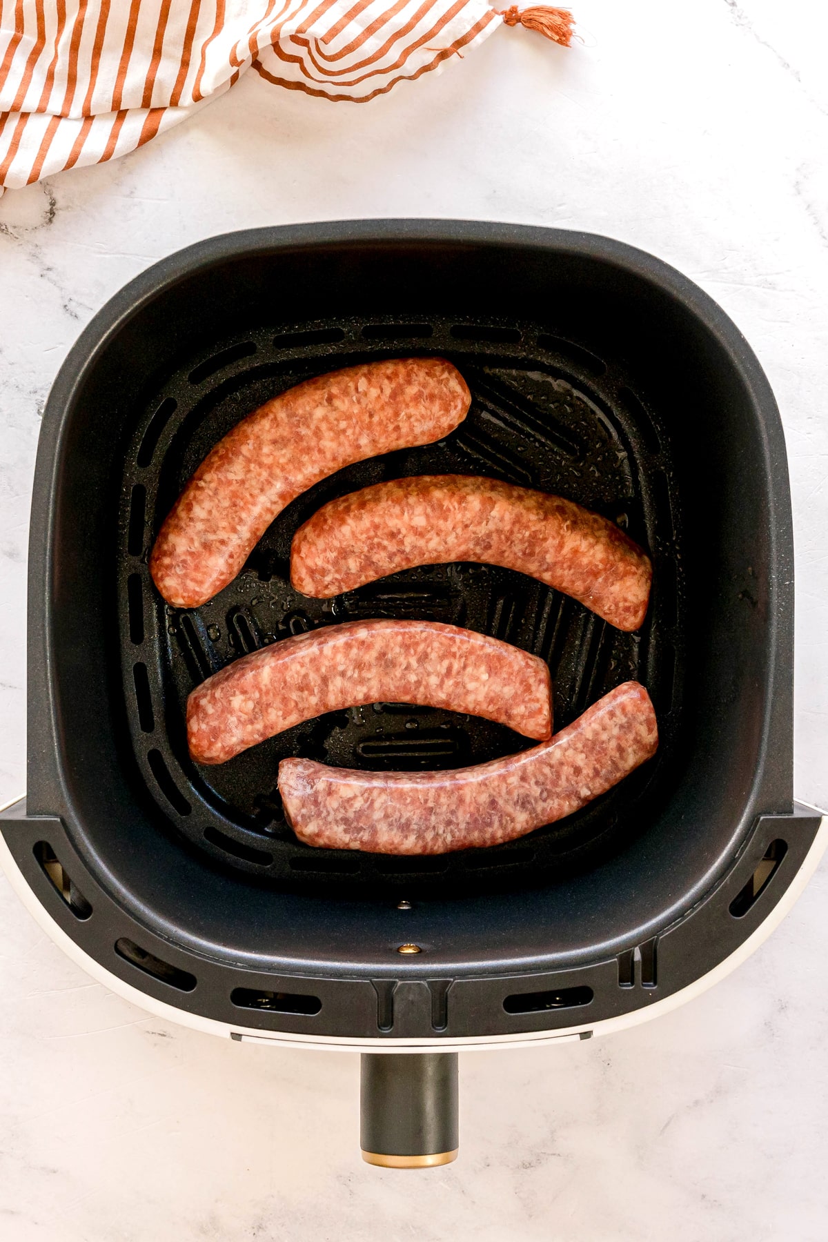 Four raw Air Fryer Brats are placed in the basket on a white surface, with a striped kitchen towel partially visible in the corner.