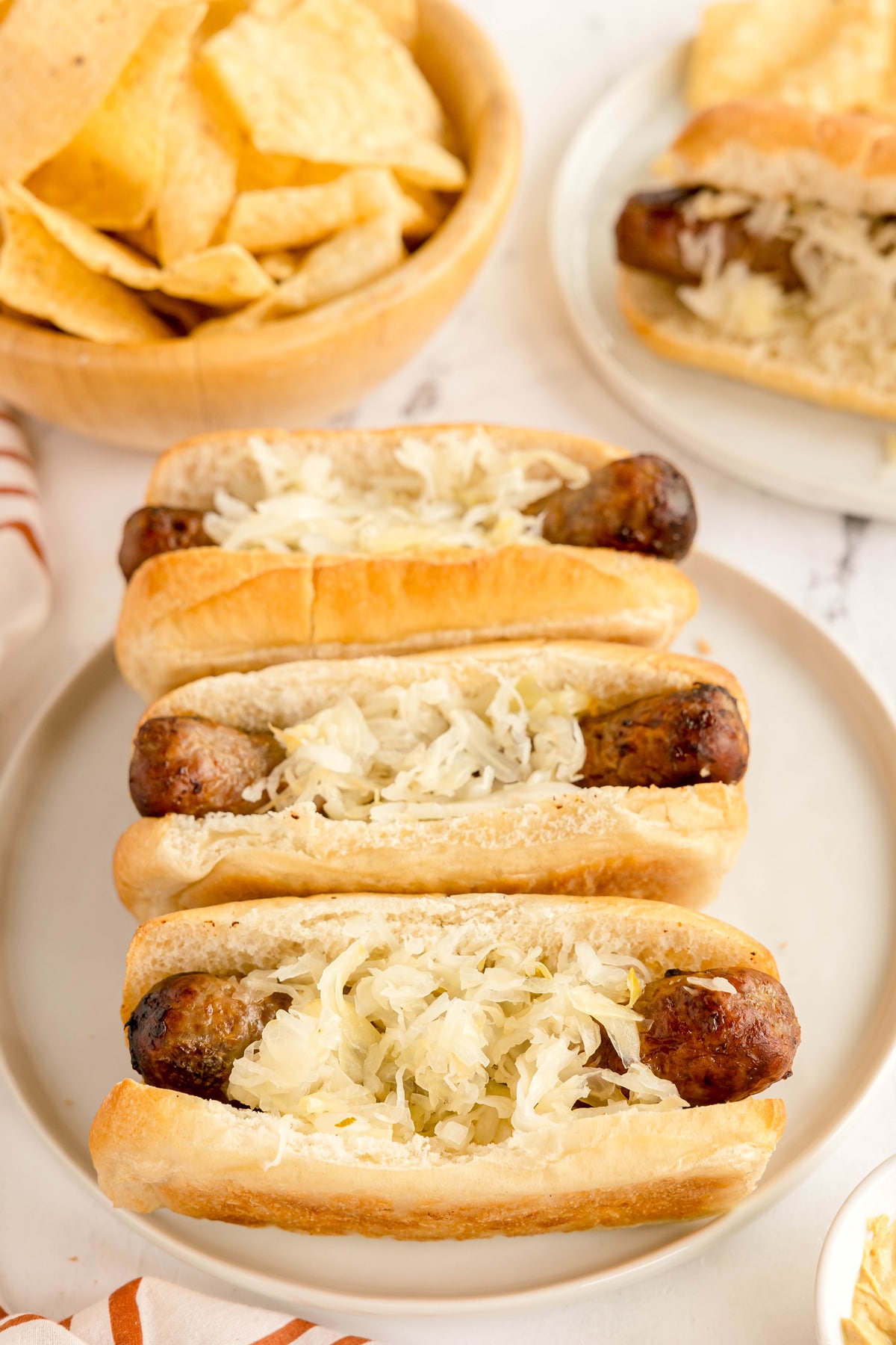 Delectable Air Fryer Brat topped with sauerkraut on a plate, with a bowl of tortilla chips and another bratwurst in the background.