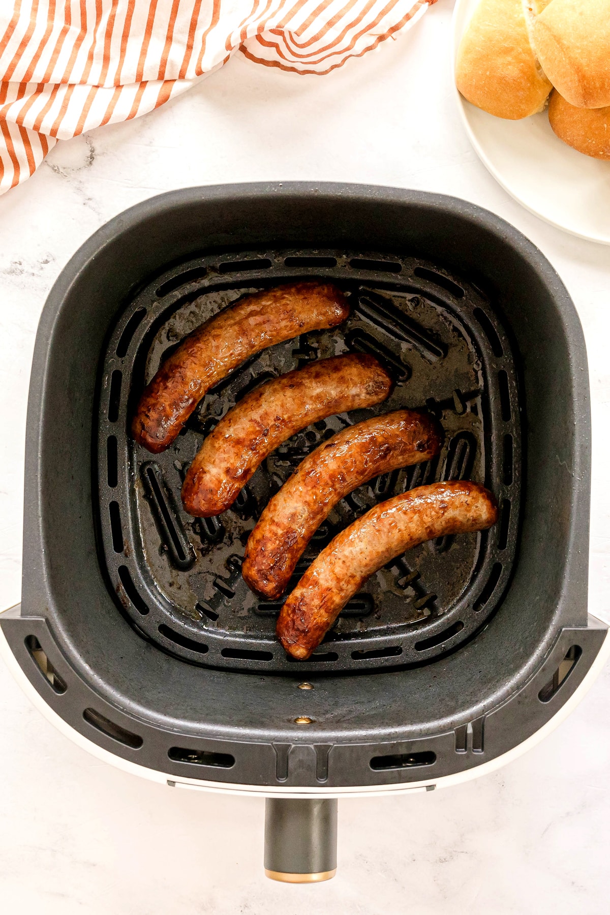 Four cooked Air Fryer Brats sit in an air fryer basket, with a plate of bread rolls and a striped cloth in the background.
