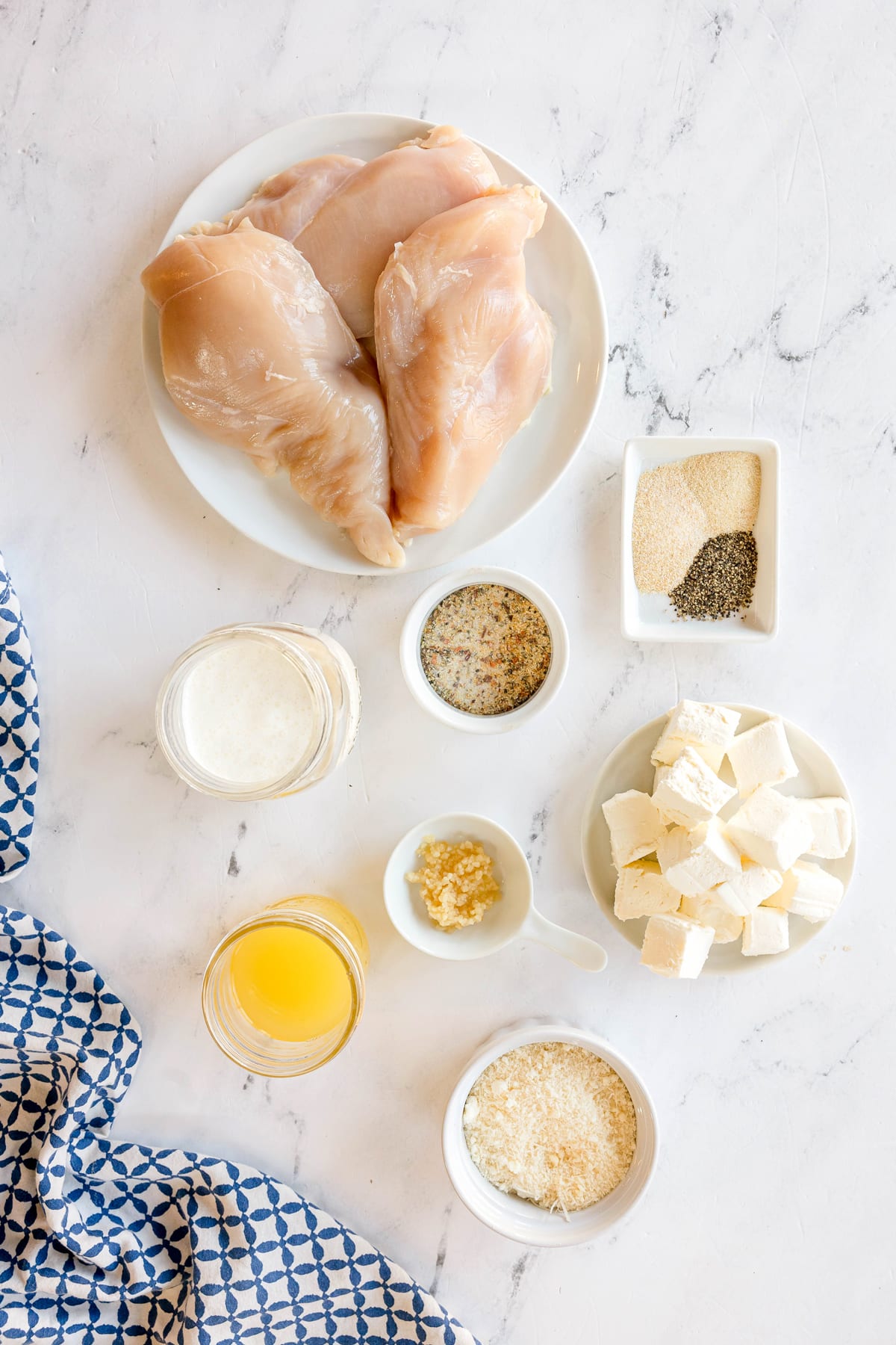 Ingredients for Slow Cooker Creamy Italian Chicken include raw skinless chicken breasts on a plate, italian seasoning mix, garlic powder, onion powder, black pepper, cream cheese, heavy cream, chicken broth, freshly grated parmesan cheese, fresh garlic cloves and parsley or basil.