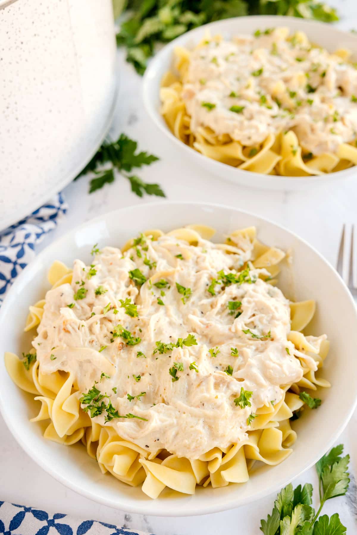 A bowl of pasta topped with Slow Cooker Creamy Italian Chicken, garnished with chopped parsley, with another similar bowl and a pot in the background.