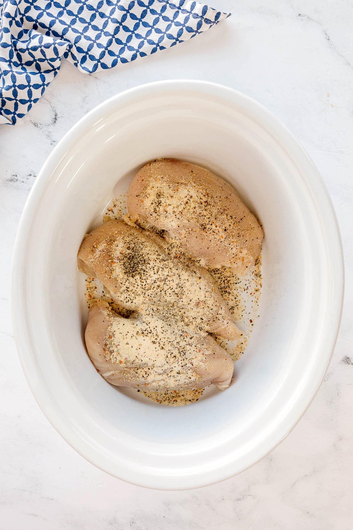 Three raw chicken breasts seasoned with Italian seasoning, garlic powder, onion powder, and pepper placed in a white slow cooker on a marble countertop, with a blue patterned towel nearby.