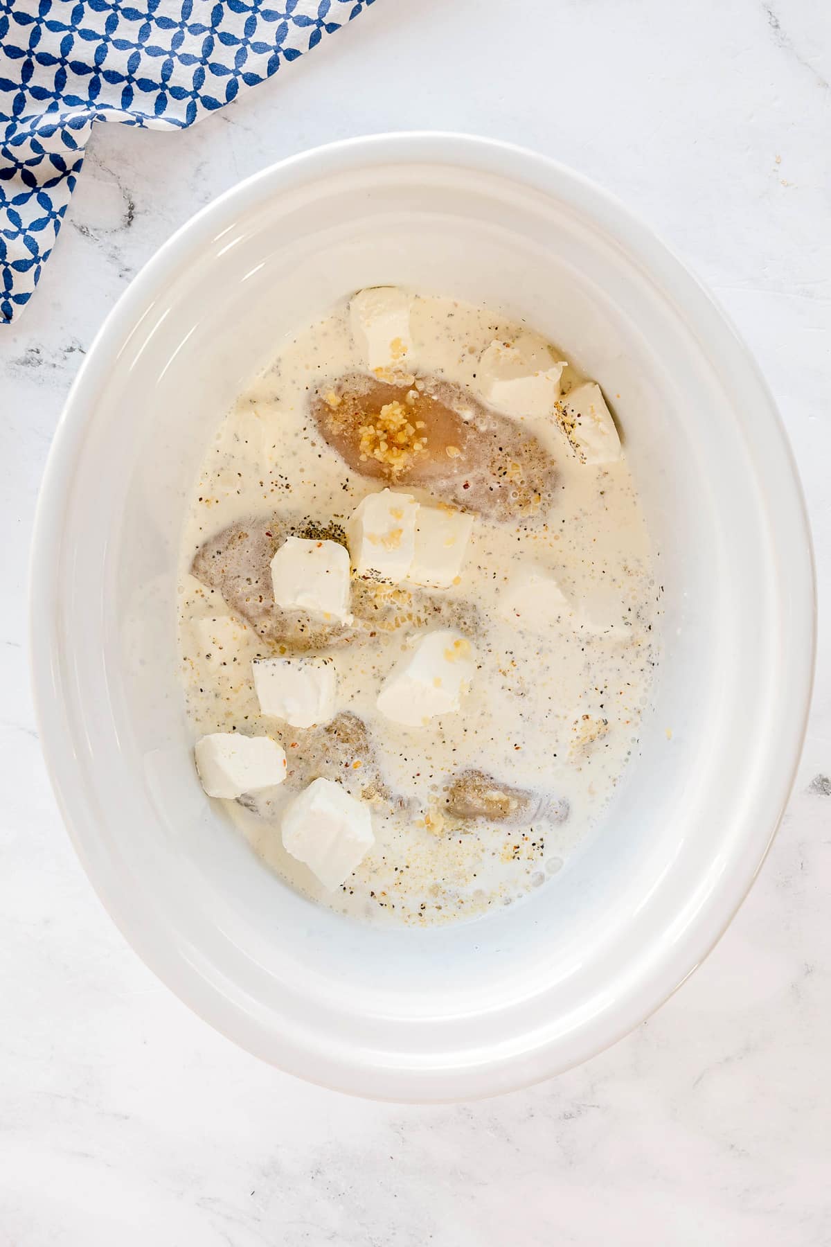 Next step in preparing Slow Cooker Creamy Italian Chicken is to add cream cheese cubes, into the mixture of minced garlic, and seasonings in a white slow cooker on a marble countertop with a blue and white cloth nearby.