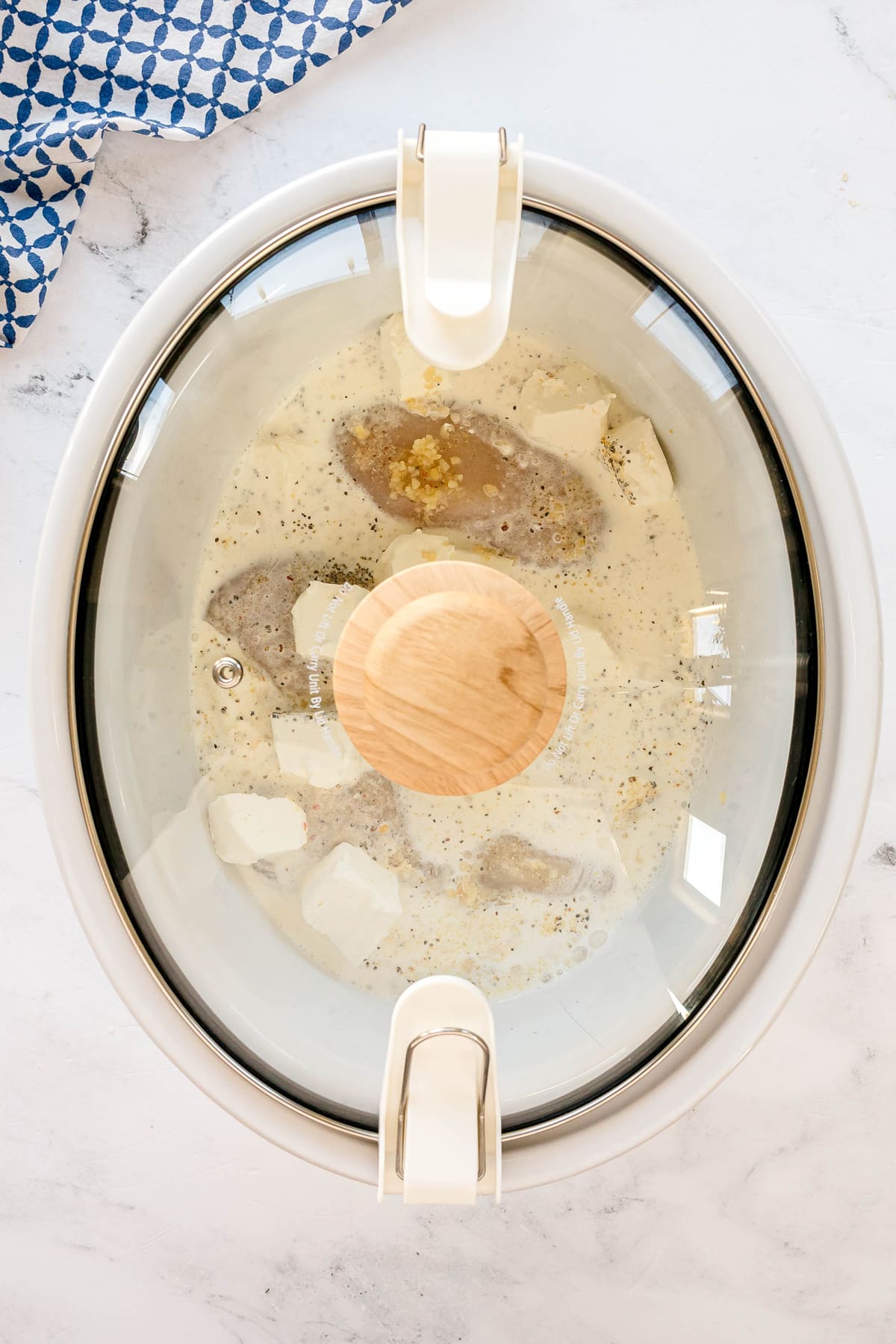 A slow cooker with a glass lid contains ingredients for Slow Cooker Creamy Italian Chicken recipe.