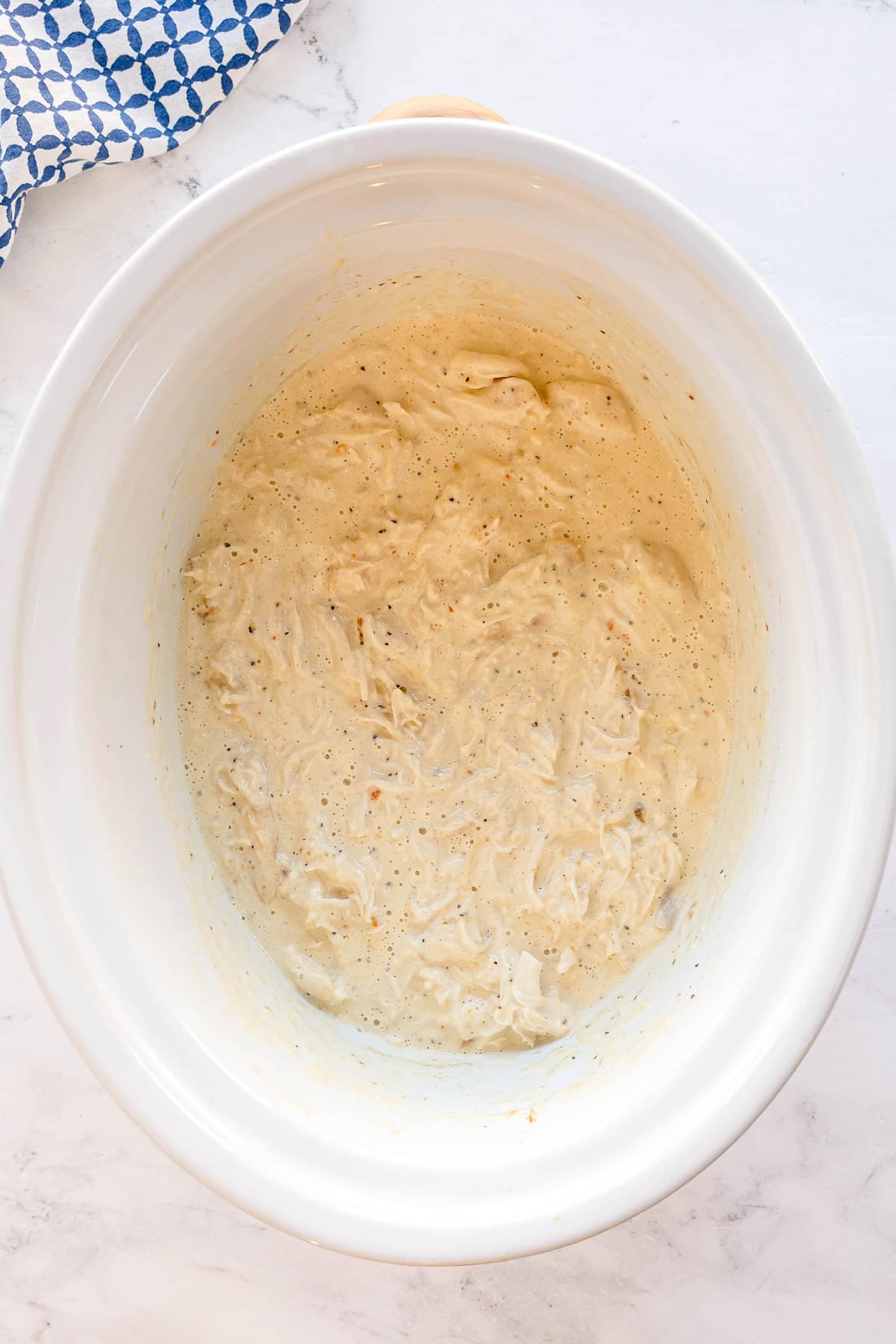 Creamy Slow Cooker Creamy Italian Chicken consisting shredded chicken is spread inside a white slow cooker on a marble countertop with a blue and white kitchen towel nearby.