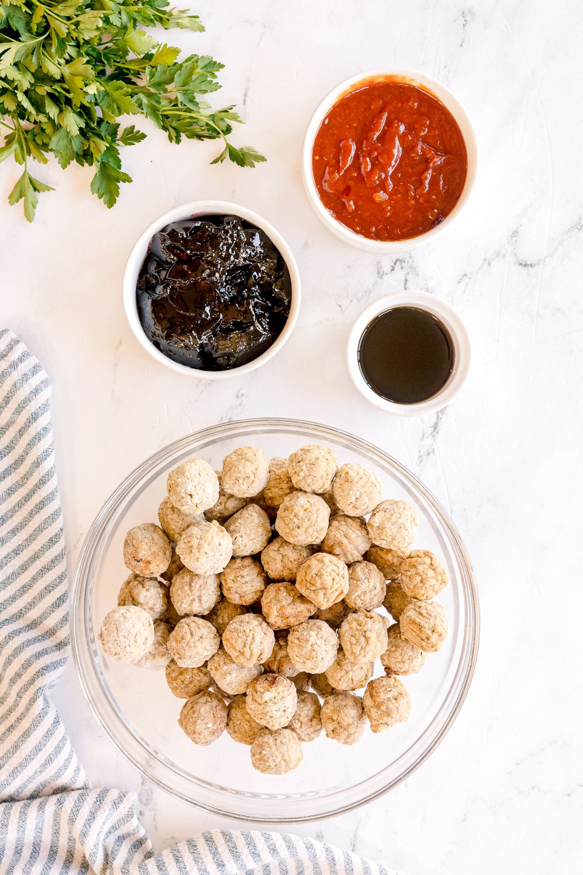 Ingredients for Meatballs with Grape Jelly and Chili Sauce in a glass bowl filled with frozen meatballs, placed next to bowls of chilli sauce, Worcestershire sauce, and grape jelly, with parsley and a striped cloth nearby on a white surface.
