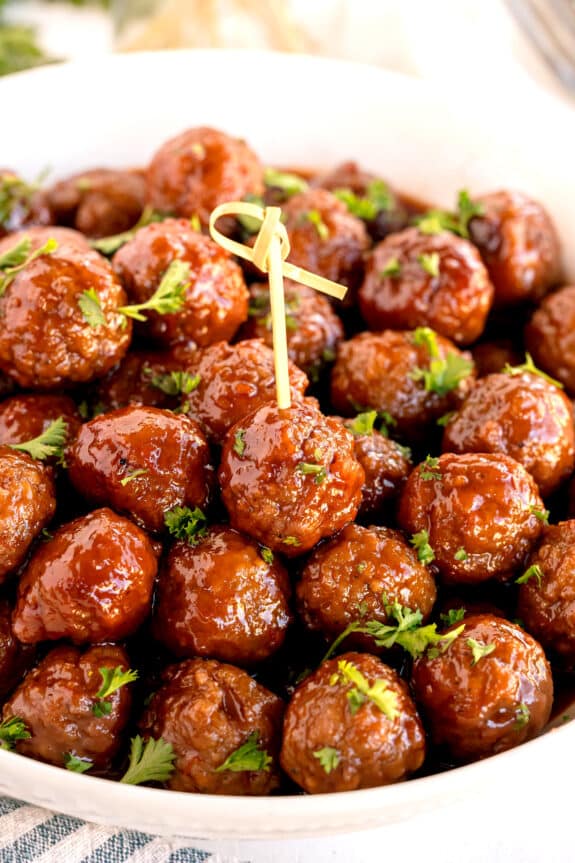 A bowl filled with Meatballs With Grape Jelly And Chili Sauce, garnished with chopped parsley, with a decorative toothpick inserted into one meatball.