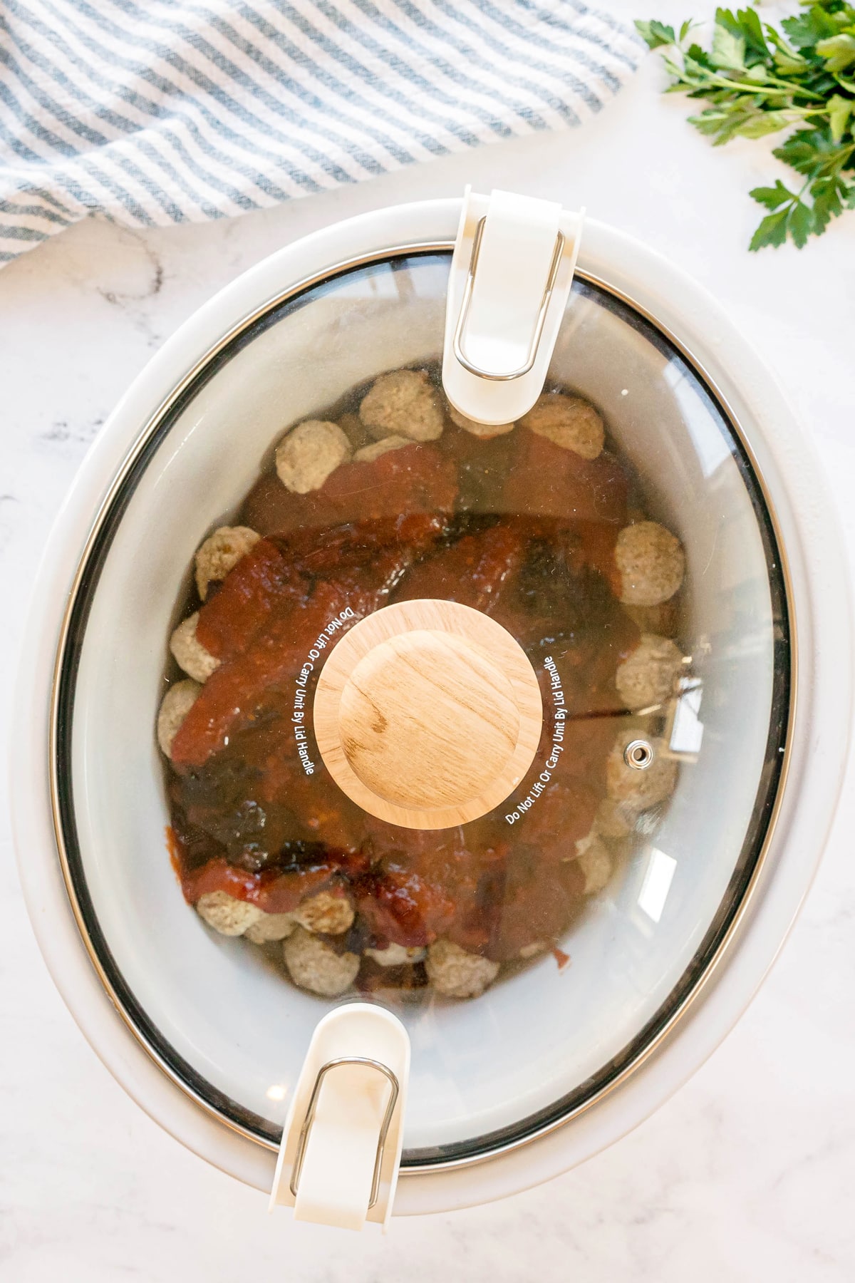 Next step in preparing Meatballs with Grape Jelly and Chili Sauce is to cover the slow cooker containing meatballs covered in sauce. A kitchen towel and fresh herbs are nearby.