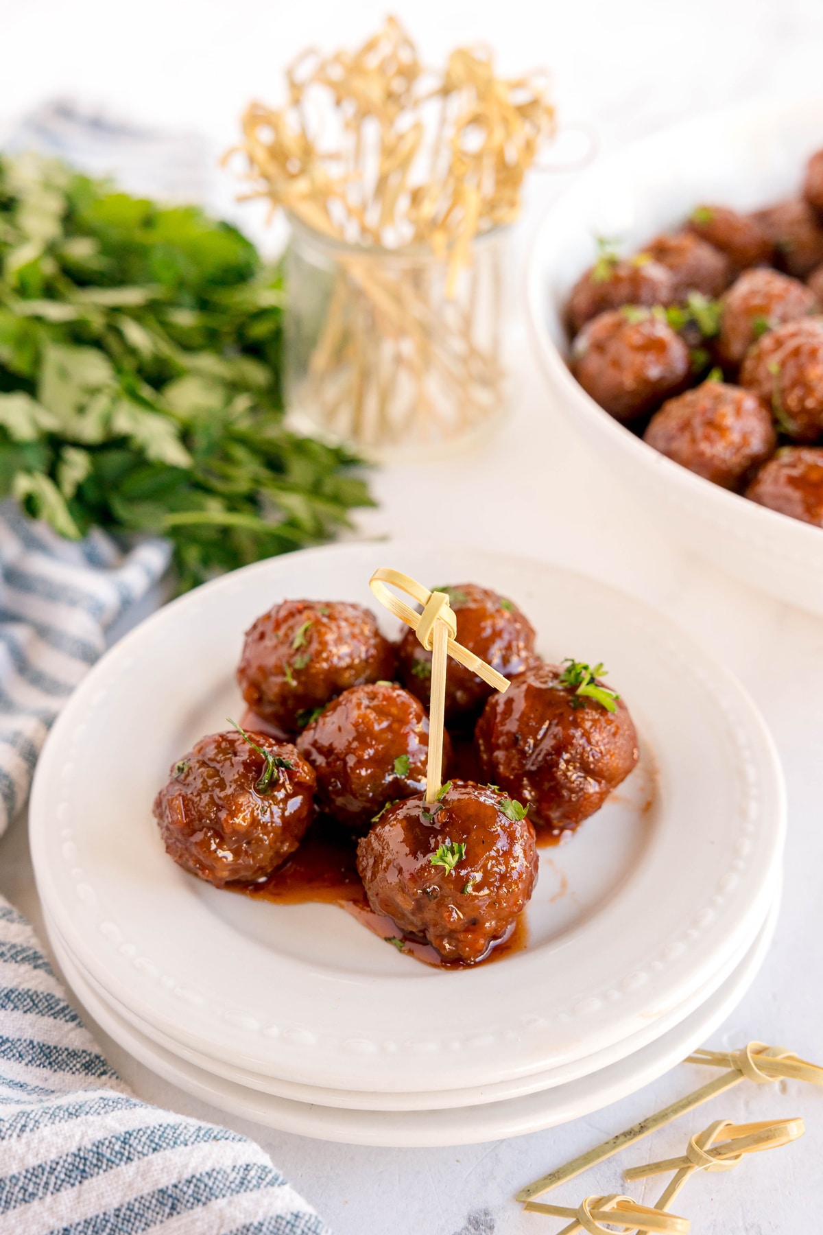 Five glazed Meatballs with Grape Jelly and Chili Sauce garnished with herbs are served on a white plate with a skewer, with more meatballs and fresh parsley in the background.