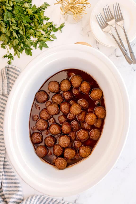 A white oval slow cooker filled with Meatballs With Grape Jelly And Chili Sauce sits on a counter next to parsley, a striped towel, forks, and white plates.