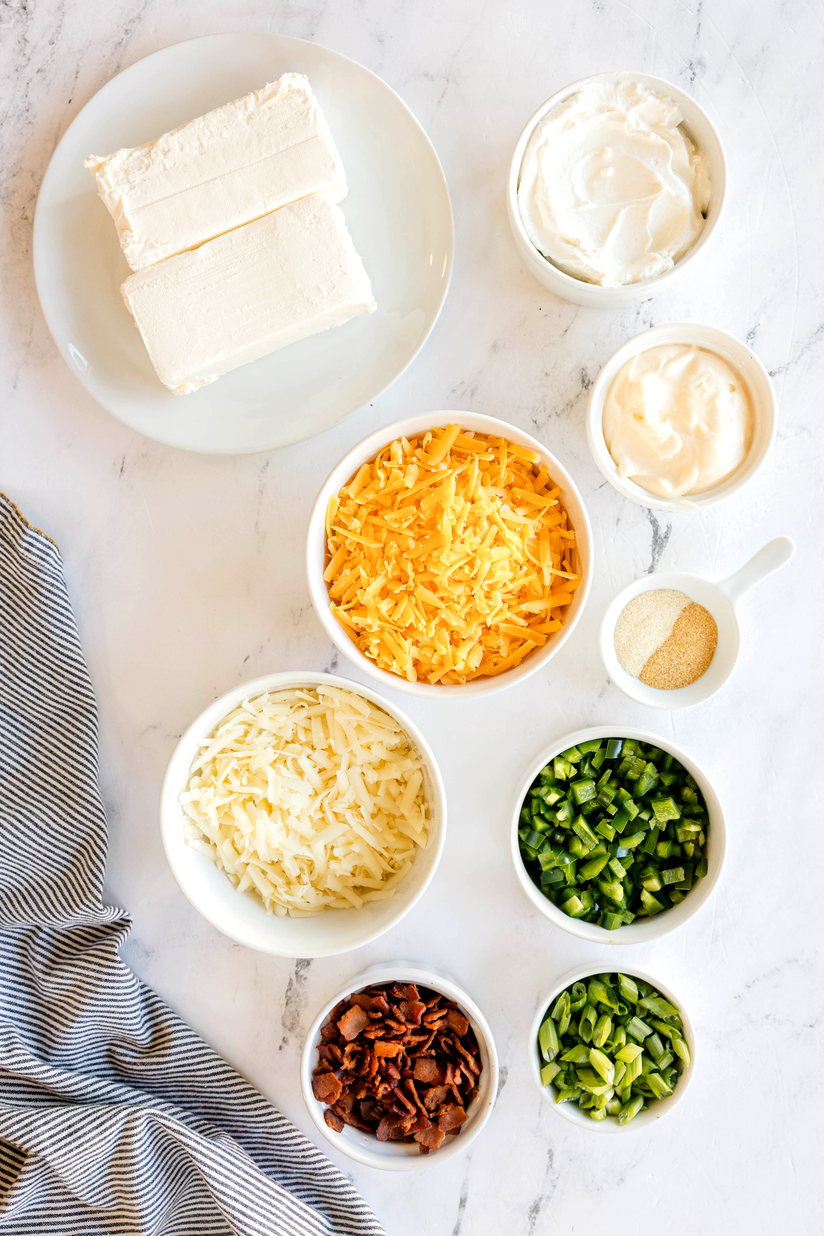 Ingredients for Crockpot Jalapeno Popper Dip, arranged in bowls on a white surface, including cream cheese, sour cream, mayonnaise, sharp cheddar cheese, Monterey Jack cheese, garlic powder, onion powder, Fresh jalapeños, bacon and green onions.