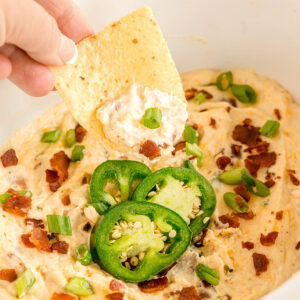 A hand dips a tortilla chip into Crockpot Jalapeno Popper Dip with sliced jalapeños, bacon bits, and chopped green onions.