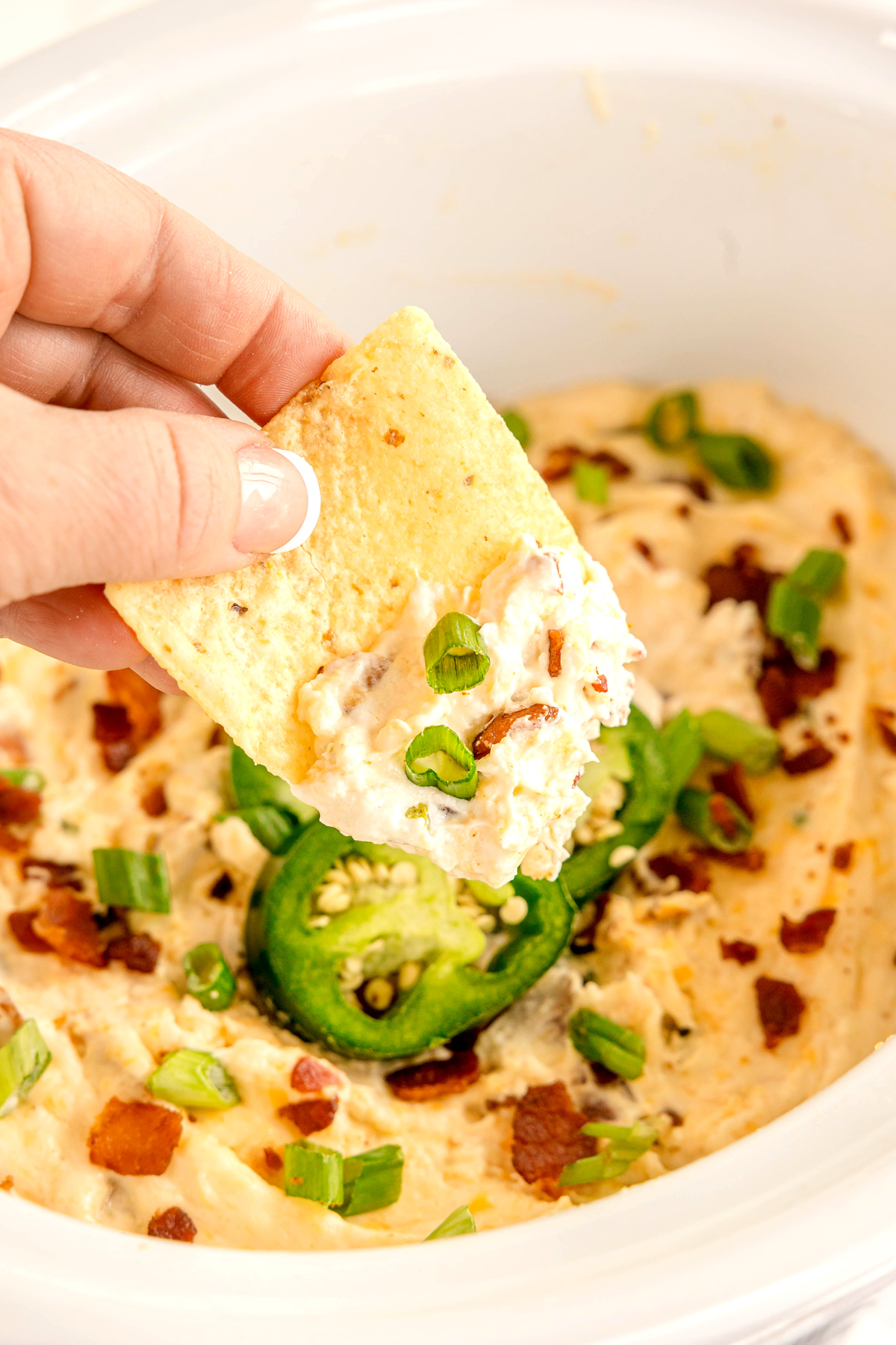 A hand holding a tortilla chip topped with creamy dip, bacon bits, chopped green onions, and sliced jalapeños or a bowl of Crockpot Jalapeno Popper Dip