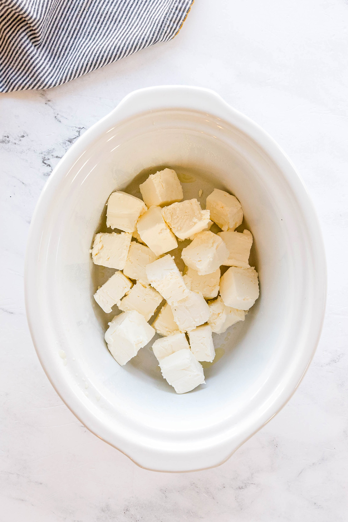 Cubes of sliced cream cheese placed in a white ceramic slow cooker on a light marble countertop with a striped cloth nearby for Crockpot Jalapeno Popper Dip recipe