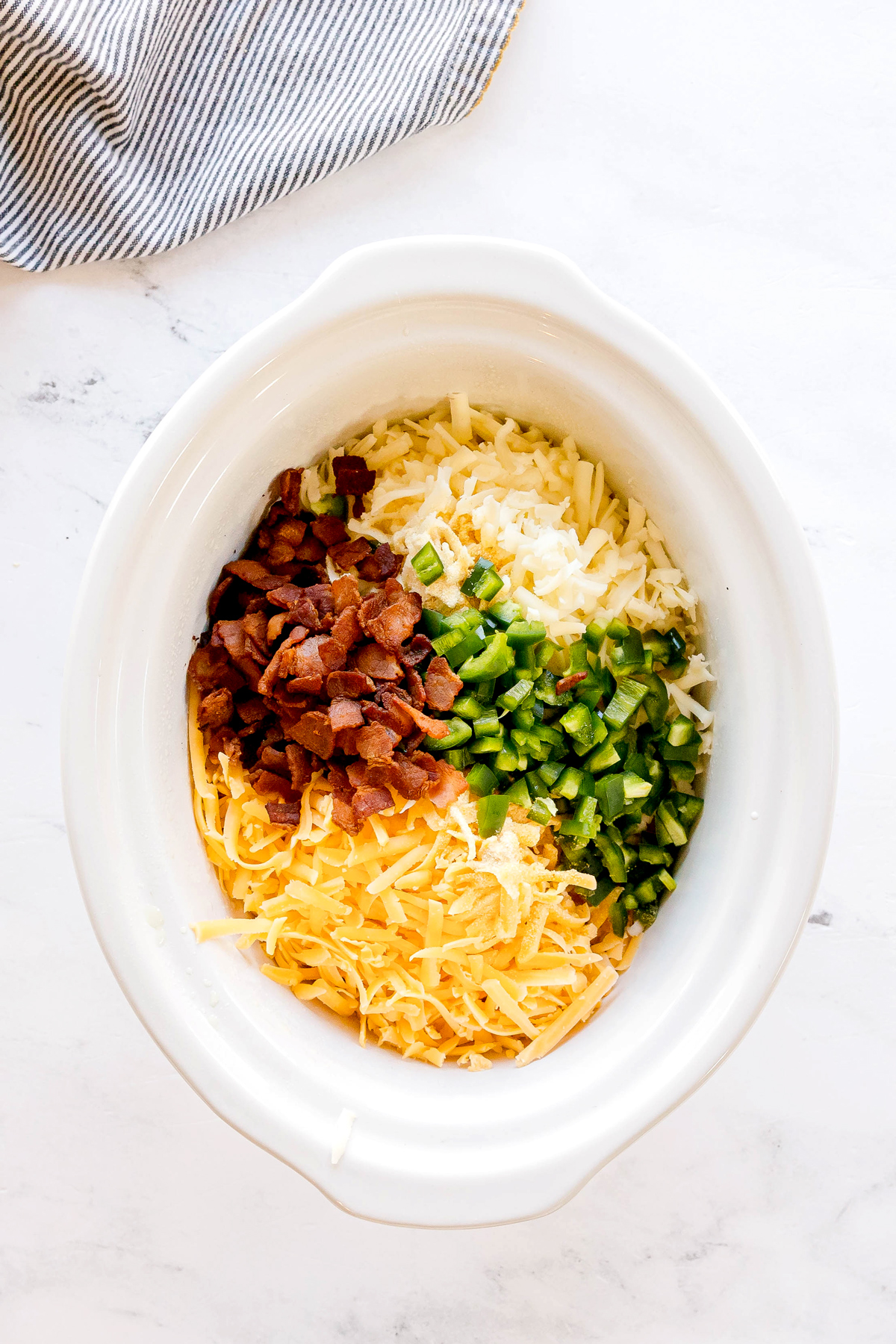 A white oval slow cooker contains sour cream, mayonnaise, both cheeses, garlic powder, onion powder, jalapeños, and half the crumbled bacon for Crockpot Jalapeno Popper Dip recipe