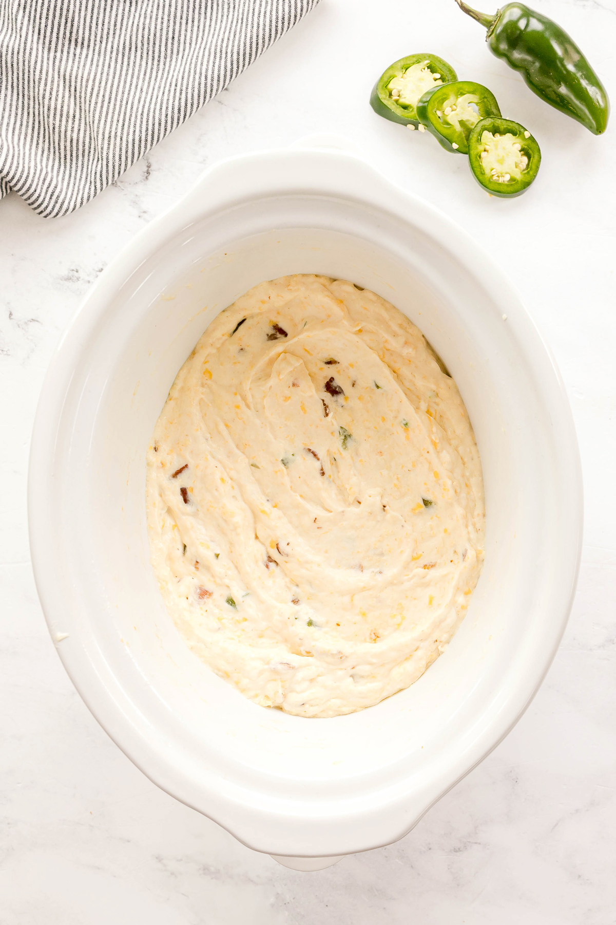 A white slow cooker filled with a cooked creamy Crockpot Jalapeno Popper Dip, next to sliced jalapeños and a striped cloth on a white countertop.