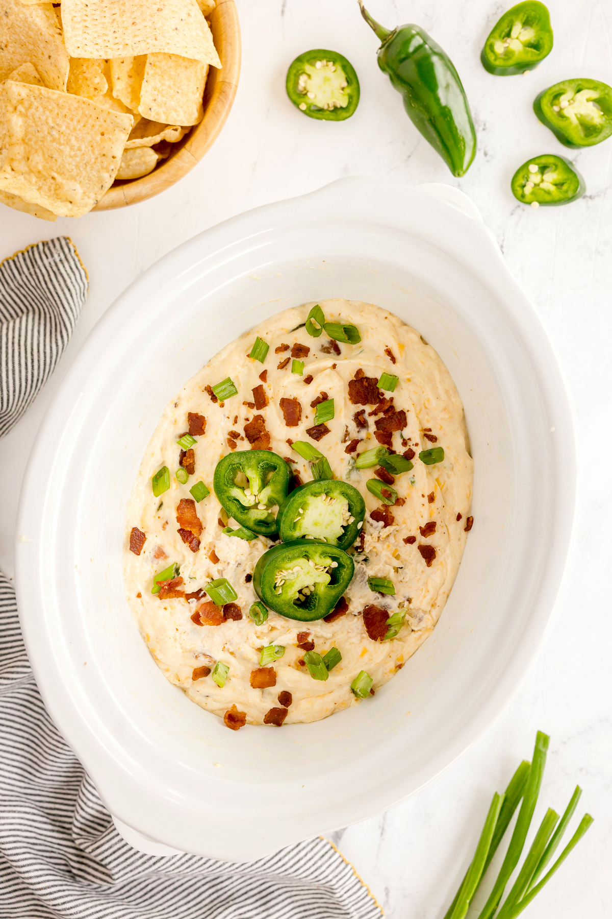 A white dish filled with Crockpot Jalapeno Popper Dip, topped with bacon bits, sliced jalapeños, and green onions; tortilla chips and jalapeños are nearby on a white surface.