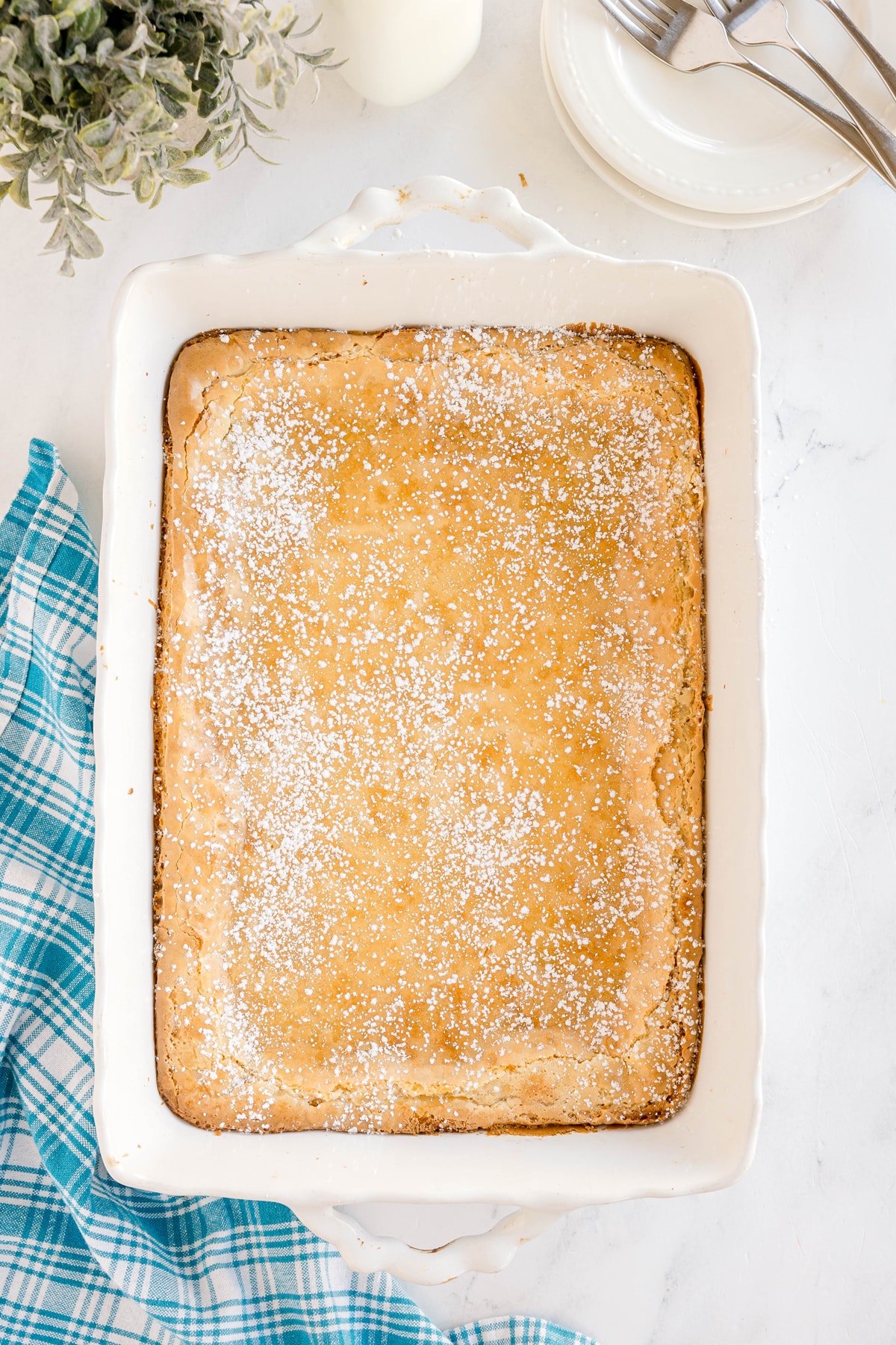 A golden-brown sheet cake dusted with powdered sugar sits in a white baking dish on a marble surface, next to a blue plaid towel and stacked plates with forks.