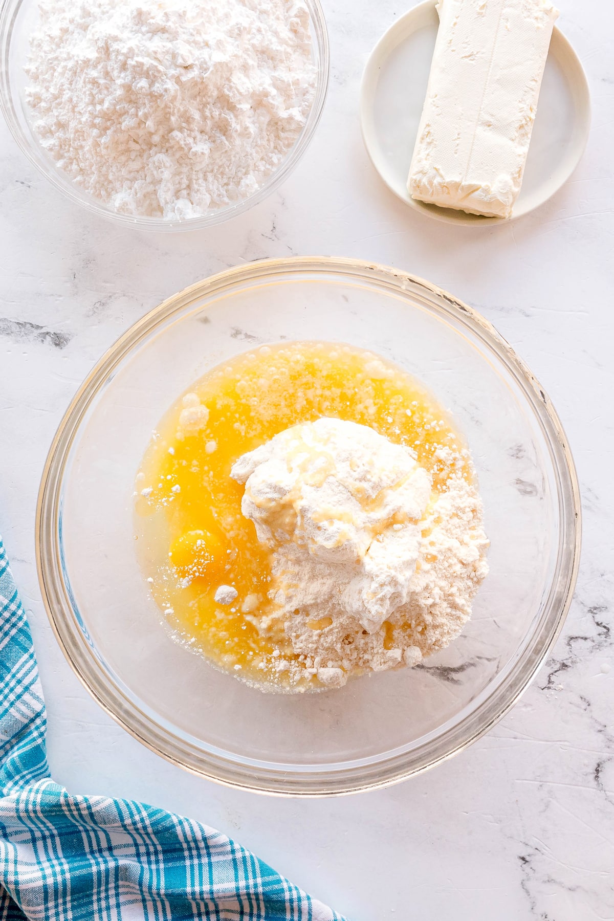 First step in preparing Gooey Cake is to use a glass bowl with flour, melted butter, eggs, and sugar sits on a white surface next to a block of cream cheese and a bowl of flour. A blue plaid cloth is nearby.