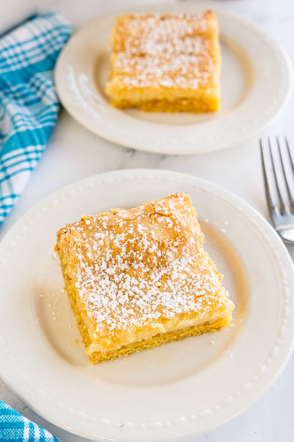 Two square pieces of Gooey Cake dusted with powdered sugar are served on white plates, with a blue checkered cloth and two forks nearby.