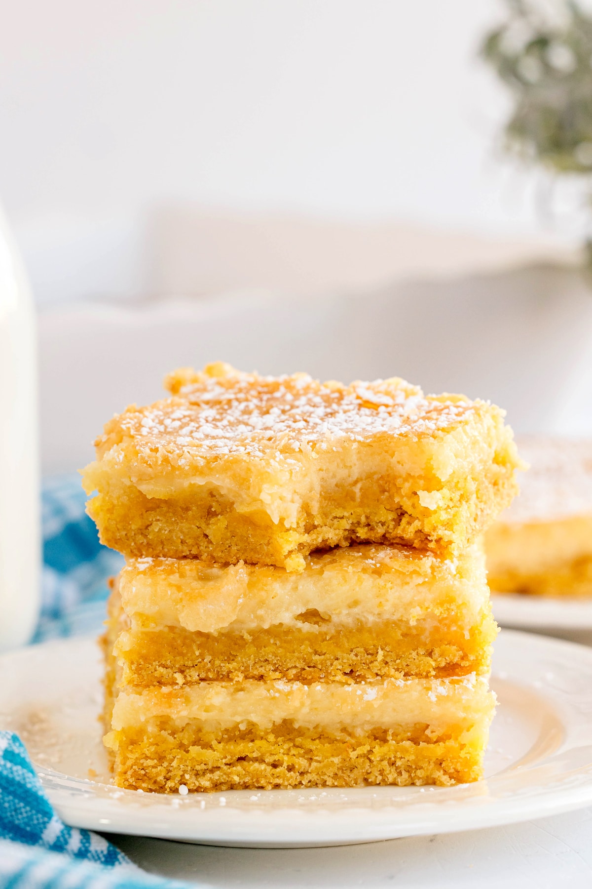 A stack of three layered lemon bars with powdered sugar on top sits on a white plate, their gooey butter cake-inspired texture promising a rich and tangy treat, with a blurred background for added allure.