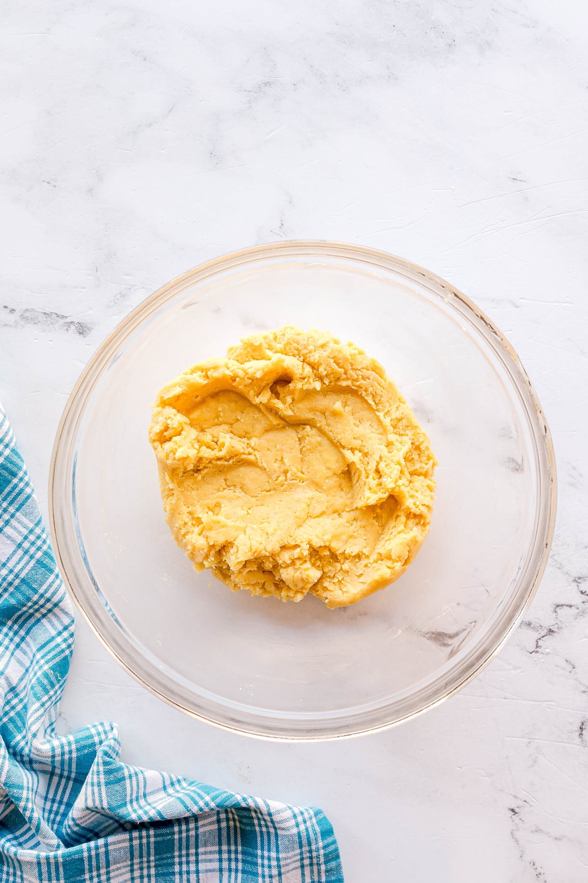 A clear glass bowl contains yellow dough for Gooey Cake on a white marble surface, with part of a blue and white plaid cloth visible on the side.