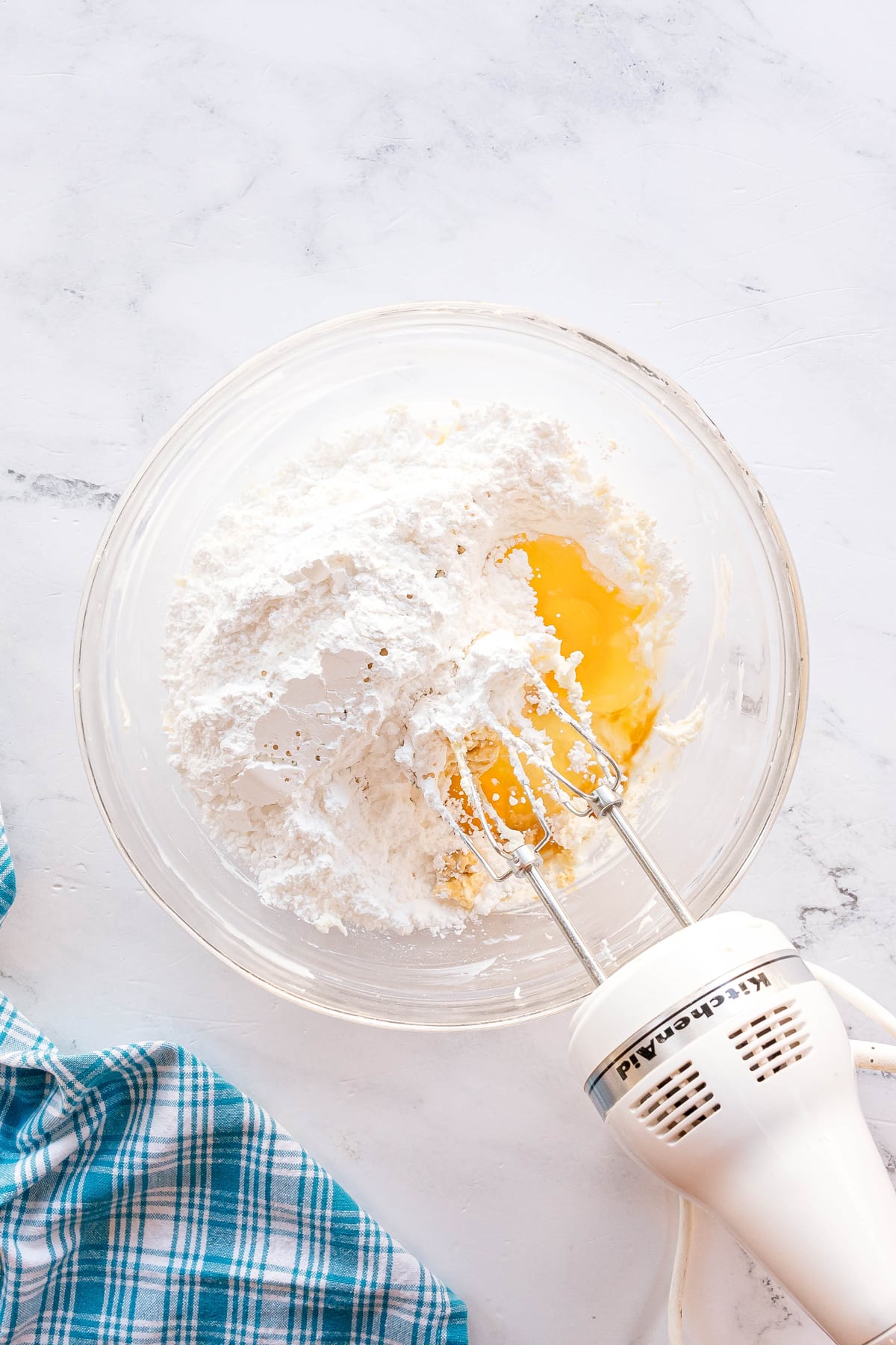 A glass bowl with flour and eggs being mixed by a hand mixer on a white countertop, next to a blue plaid kitchen towel.