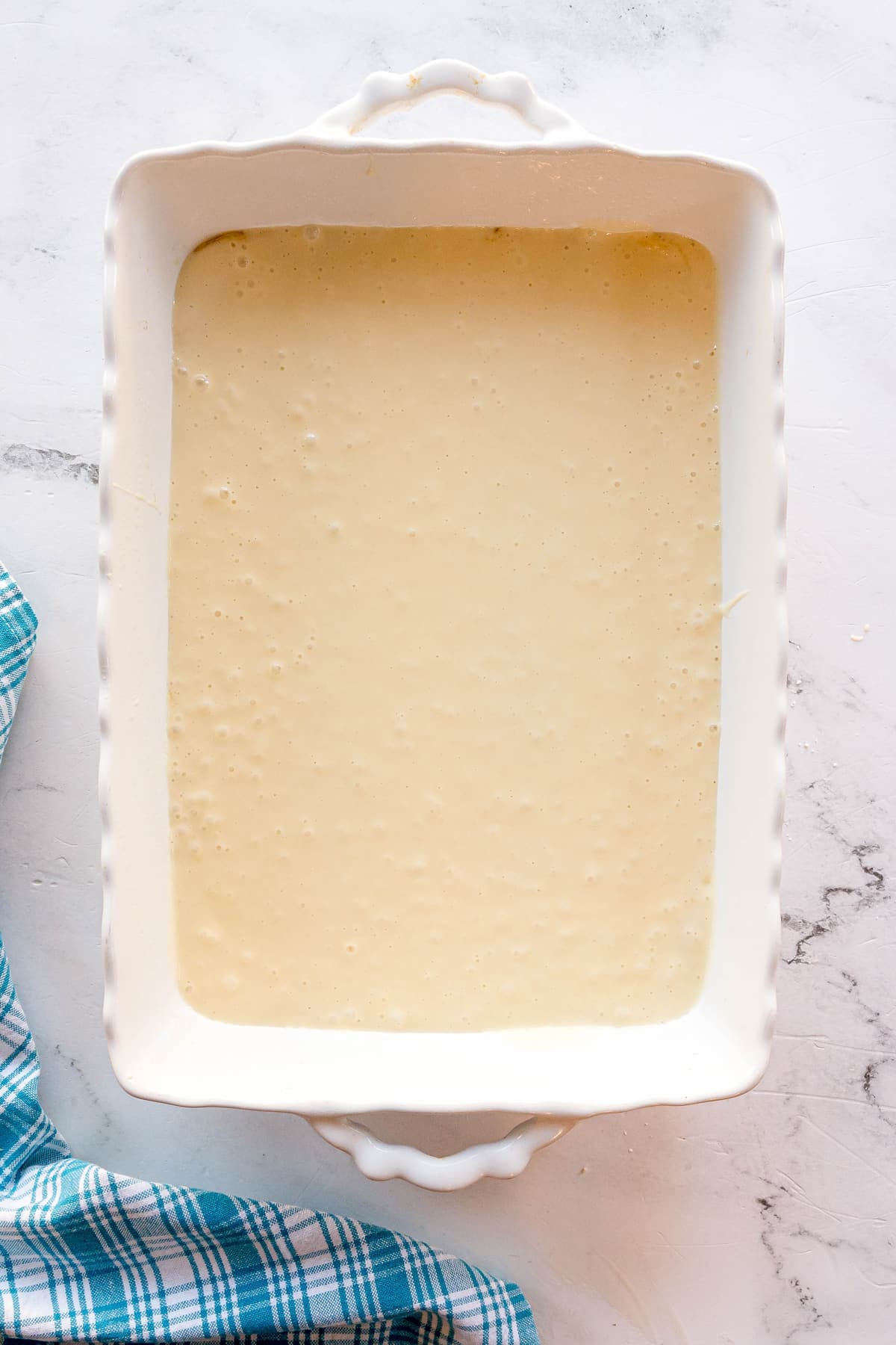 A rectangular white baking dish filled with a pale yellow liquid batter sits on a white surface next to a blue and white checkered cloth.