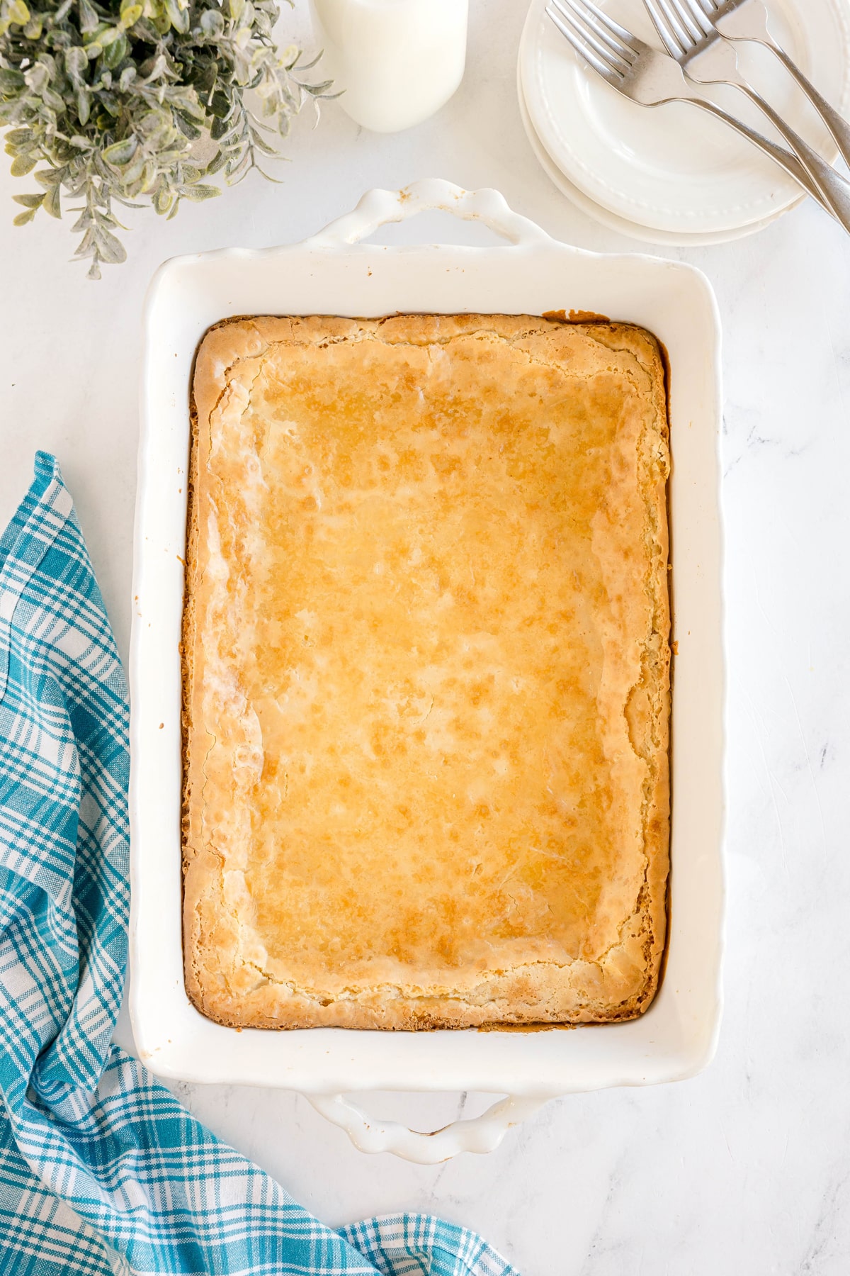 A golden-brown baked dessert in a rectangular white dish sits on a marble surface beside a blue plaid cloth, a plant, a bottle of milk, and stacked plates with forks.