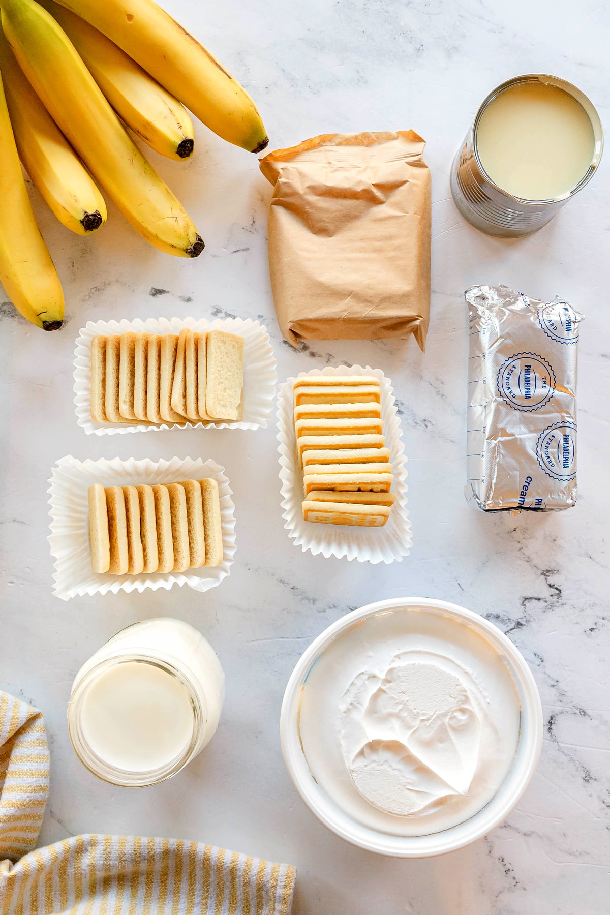 Ingredients for Paula Deen Banana Pudding arranged on a marble surface, include: frozen whipped topping, sweetened condensed milk, cream cheese, milk, instant french vanilla pudding mix, bananas, and Chessmen cookies.