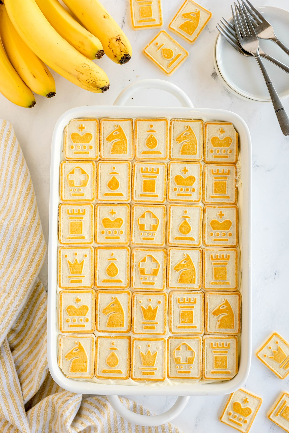A rectangular dish filled with a Paula Deen Banana Pudding with square chess-themed cookies, next to bananas, forks, and a striped cloth.