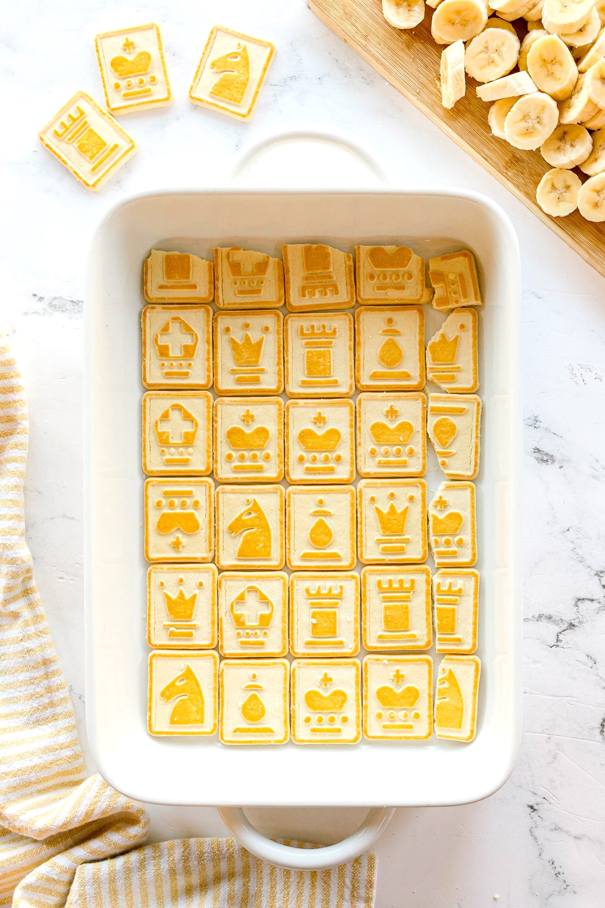 A white baking dish filled with Chessmen cookies designs. Sliced bananas are on a cutting board nearby.