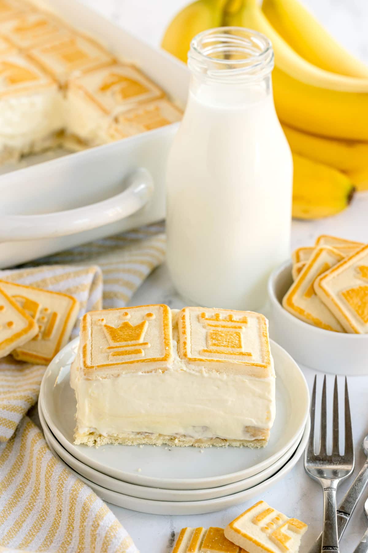 A plate with a slice of Paula Deen Banana Pudding topped with square cookies, next to a bottle of milk, a fork, and bananas in the background.