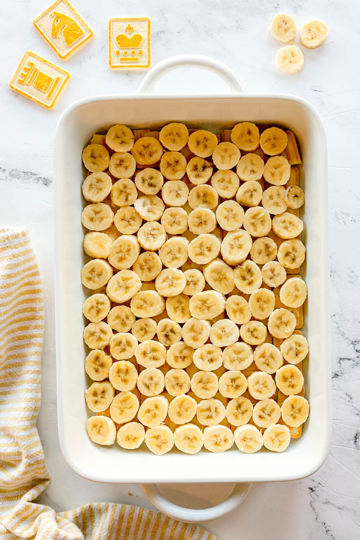 This is the next step in preparing Paula Deen Banana Pudding. Adding evenly sliced bananas arranged in rows on a marble surface, with decorative cookies and a striped towel nearby.