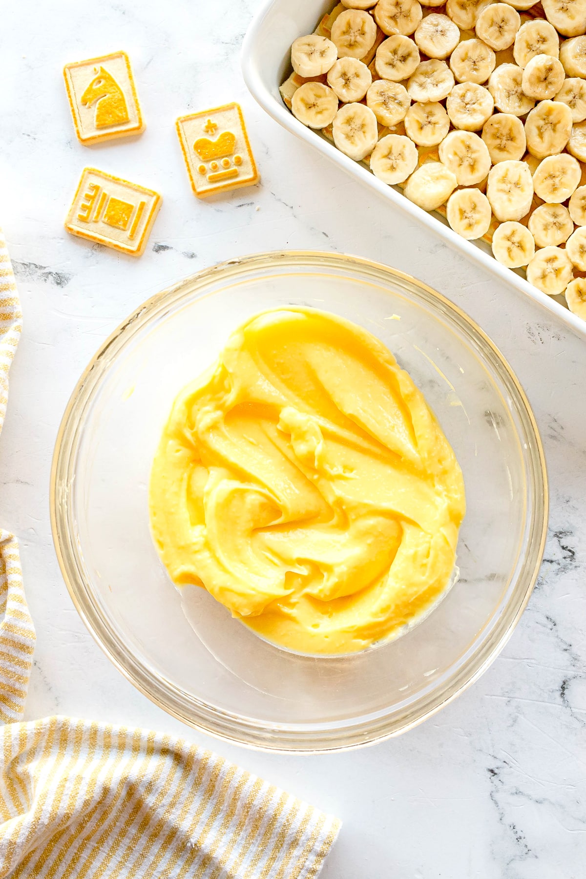 Next step in preparing Paula Deen Banana Pudding is to mix milk and pudding mix, resuting to a glass bowl of yellow pudding. A baking dish filled with sliced bananas, and chess-themed cookies on a white surface.