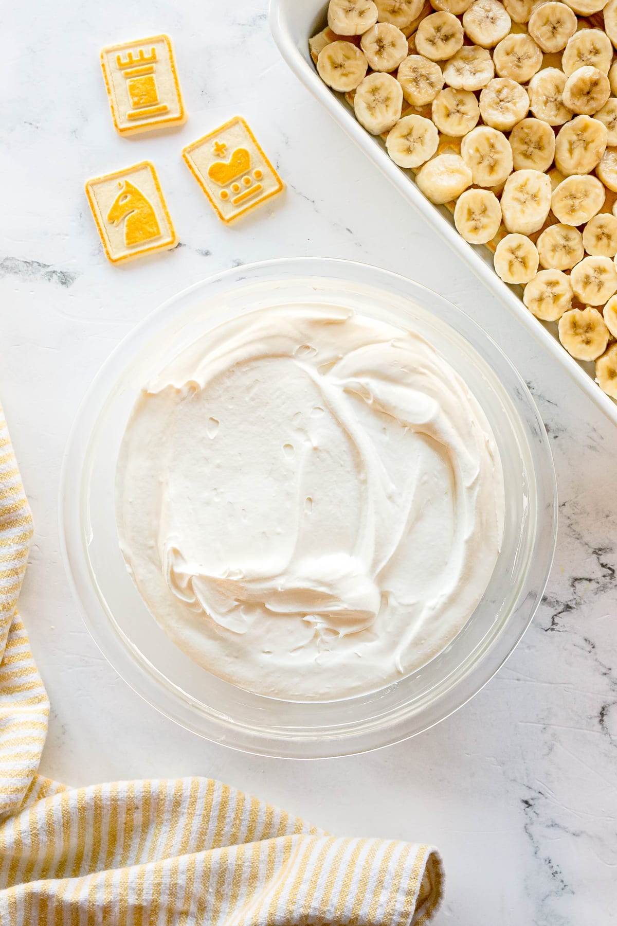 A bowl of whipped cream for Paula Deen Banana Pudding recipe sits next to a dish of sliced bananas, chess-themed cookies, and a yellow-striped towel on a marble surface.