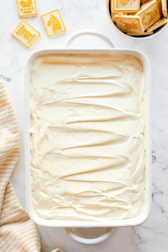 A rectangular white baking dish filled with smooth, swirled frosting sits on a marble surface next to letter and animal cookies and a striped towel. This is one step in preparing Paula Deen Banana Pudding