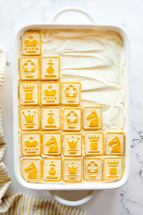 A rectangular baking dish of Paula Deen Banana Pudding topped with chess-themed cookies arranged in neat rows, covering about half of the dessert.