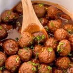 A wooden spoon stirs a pot of Meatballs With Grape Jelly And Chili Sauce, garnished with chopped parsley in a rich brown glaze.
