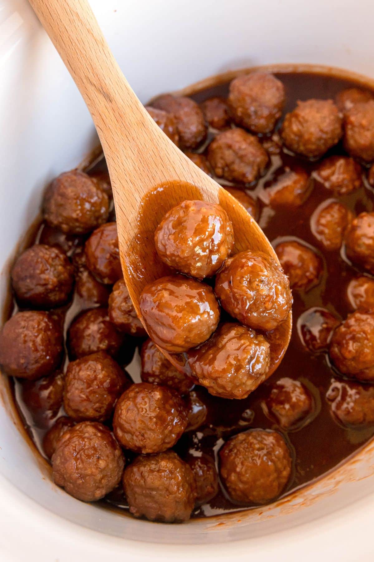 A wooden spoon holds saucy Meatballs With Grape Jelly And Chili Sauce above a slow cooker filled with more meatballs in a thick, dark brown sauce.