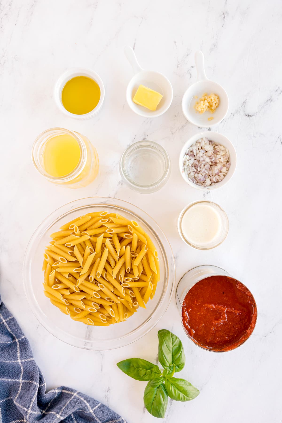 Top-down view of Pasta Alla Vodka ingredients include uncooked penne pasta, crushed tomatoes, cream, butter, garlic, shallots, vodka, chicken stock, salt and pepper, heavy cream, freshg basil, and crusty bread