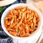A bowl of Pasta Alla Vodkain a creamy tomato sauce, topped with grated cheese and chopped basil, placed on a table with a slice of bread nearby.