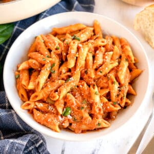 A bowl of Pasta Alla Vodkain a creamy tomato sauce, topped with grated cheese and chopped basil, placed on a table with a slice of bread nearby.
